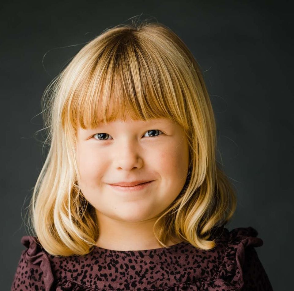 Nærbillede af glad lille pige med lyst hår, der smiler venligt til et børnefoto i naturligt lys.