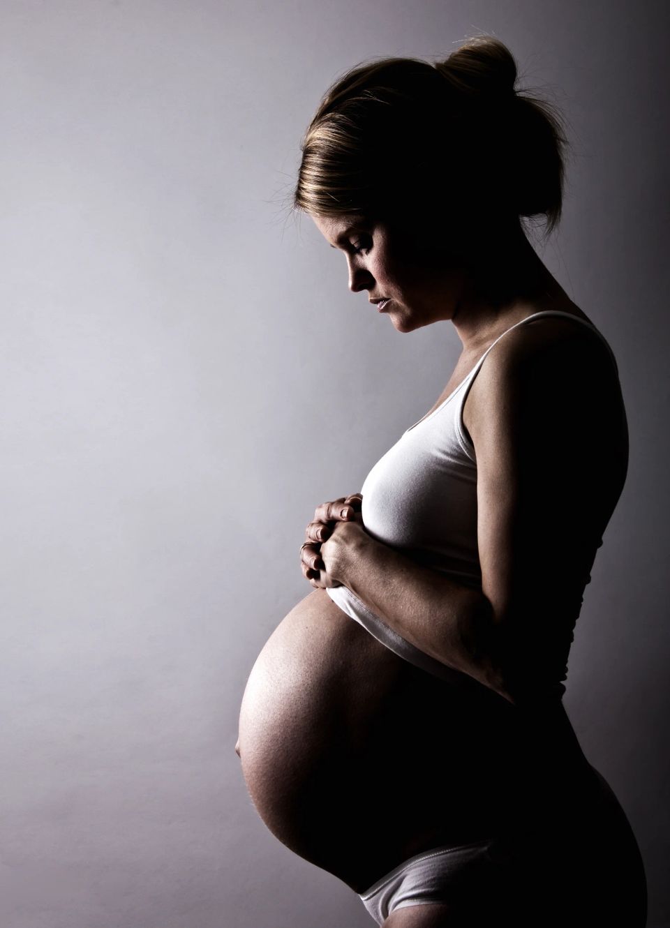 Gravid kvinde holder sin mave i profil, badet i blødt sidelys. Kontrastfuldt graviditetsfoto.