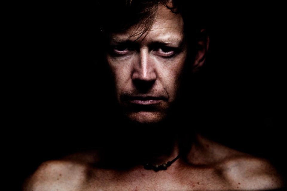 Intensivt portrætfoto af mand med rynker, oplyst af skarpt lys fra oven mod mørk baggrund, skaber dramatiske skygger.