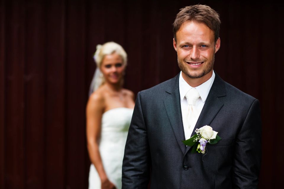 Bryllupsfoto med smilende brudgom i fokus, bruden sløret i baggrunden. Varmt lys og glad stemning.