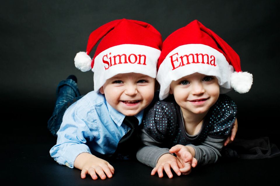 Børnefoto af to glade, smilende børn i julehuer med navne, liggende på maven mod mørk baggrund.