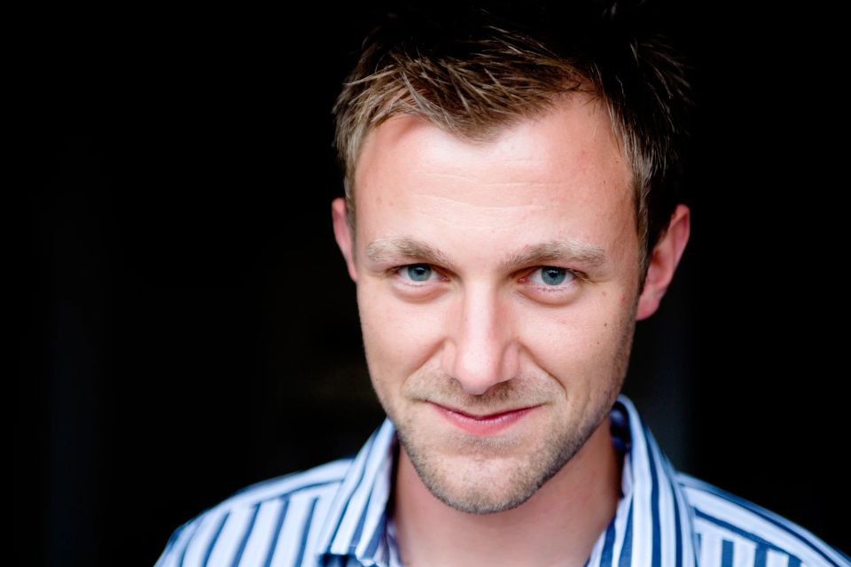 Nært portrætfoto af en smilende mand med lyst hår og intense blå øjne, kigger direkte ind i kameraet.