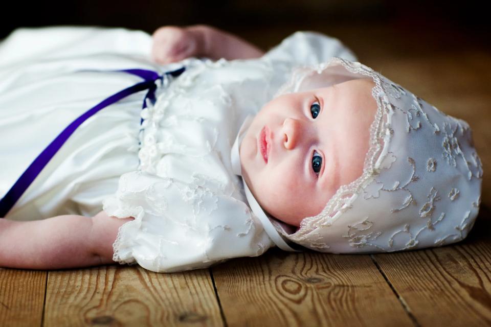 Nærbillede af baby liggende på trægulv, iført hvid kjole og kyse, med store blå øjne, et babyfoto.