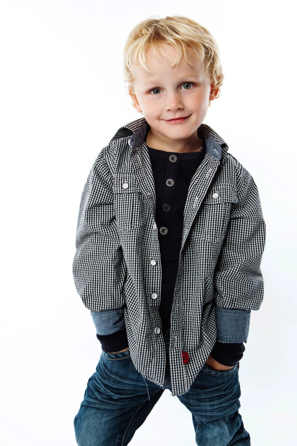 Glad lille dreng med blond hår i ternet skjorte og jeans, stående mod hvid baggrund. Et charmerende børneportræt.