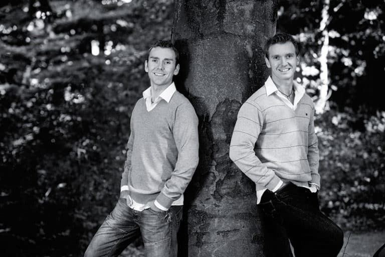 Sort/hvid portrætfoto af to smilende brødre i Virum, der læner sig afslappet op ad et træ i blødt lys.
