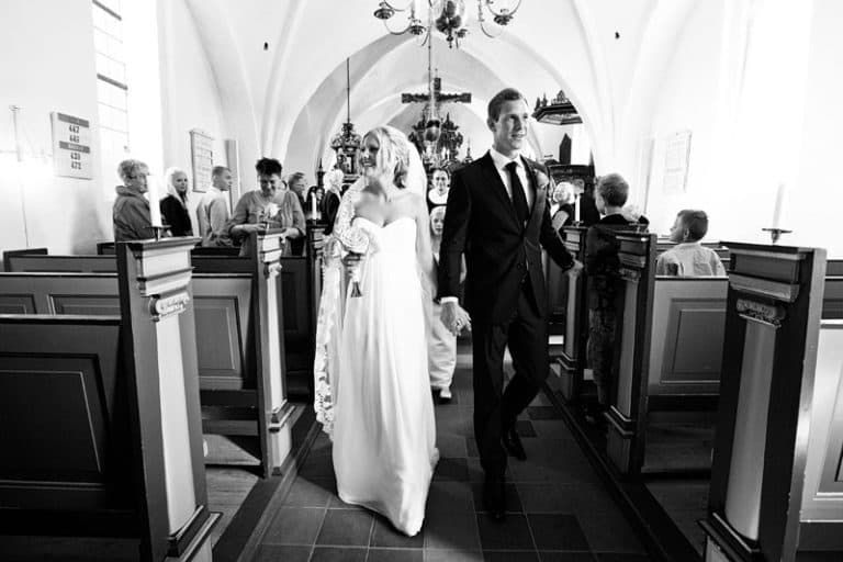 Bryllupsfoto af smilende brudepar hånd i hånd på vej ud af kirken efter vielsen i dagslys.