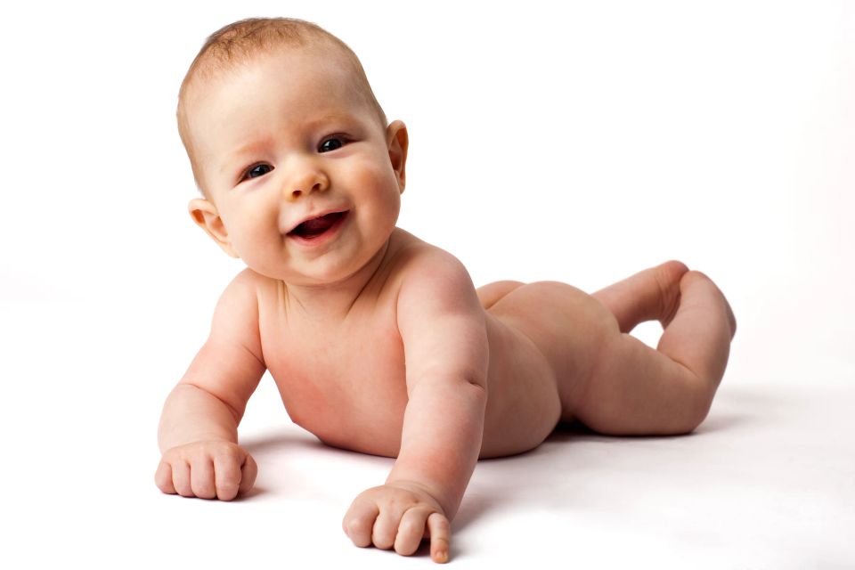 Glad baby ligger på maven på hvid baggrund, kigger direkte på kameraet med et stort smil. Et dejligt babyfoto.