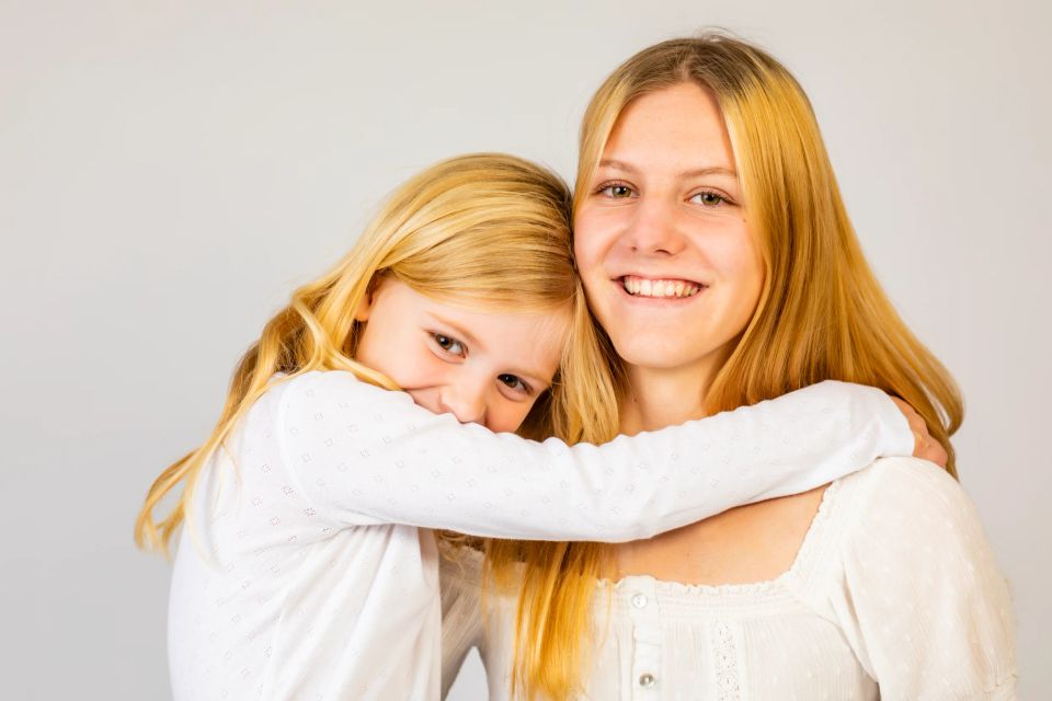 Familiebillede af to søstre med lyst hår, der omfavner hinanden og smiler i naturligt lyst studie fotografering
