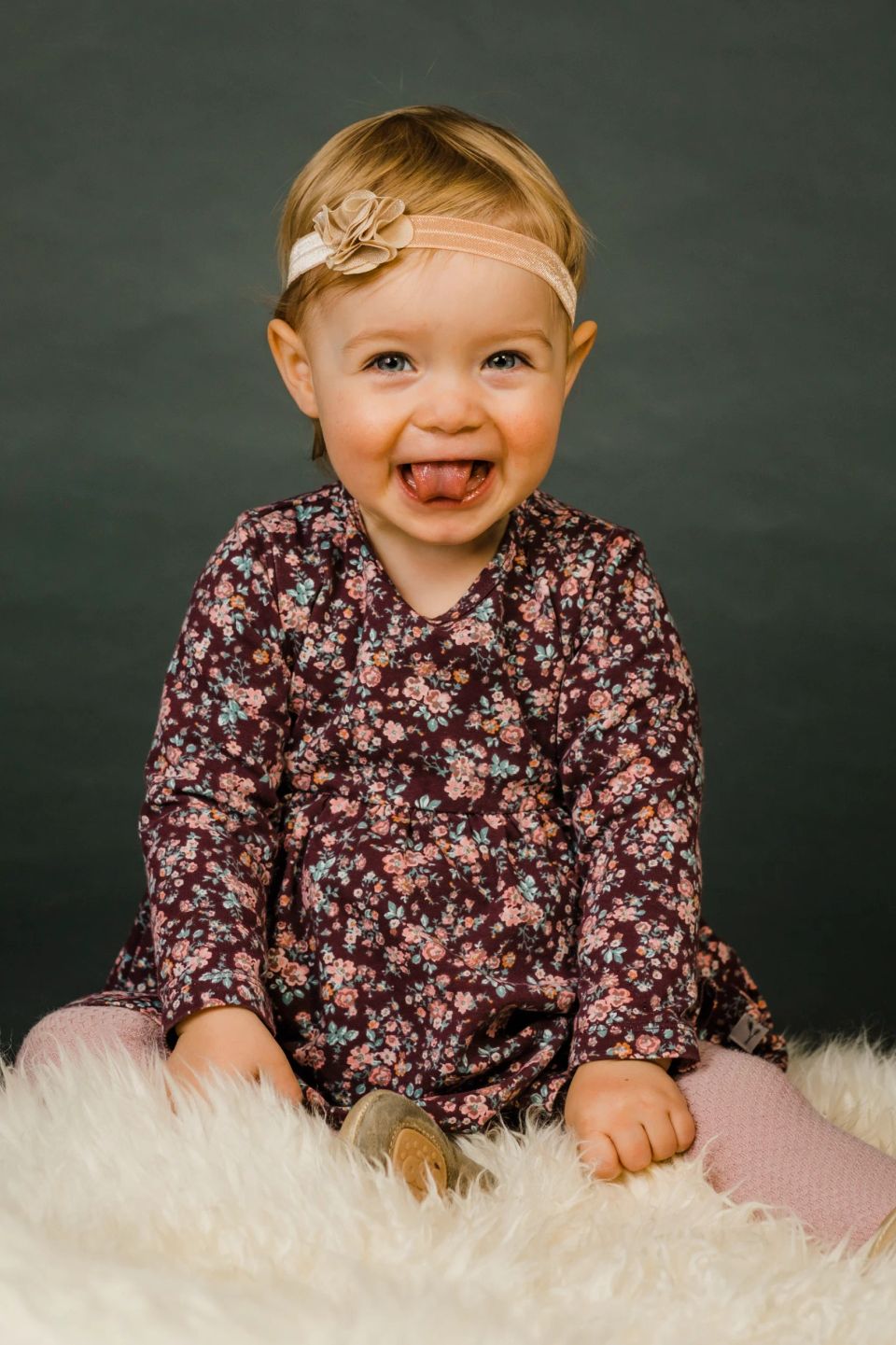 Glad barn i blomstret kjole og pandebånd sidder på hvidt lammeskind, rækker tunge mod kameraet i Virum.