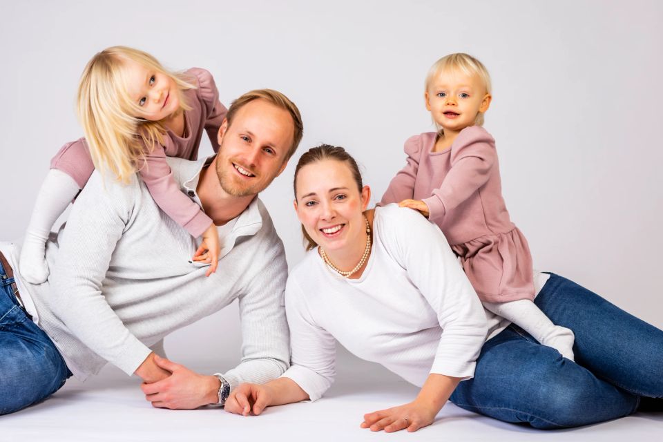 Familiefoto med mor, far og to små piger liggende og smilende på gulvet i legesyg stemning. Lyst, venligt lys.