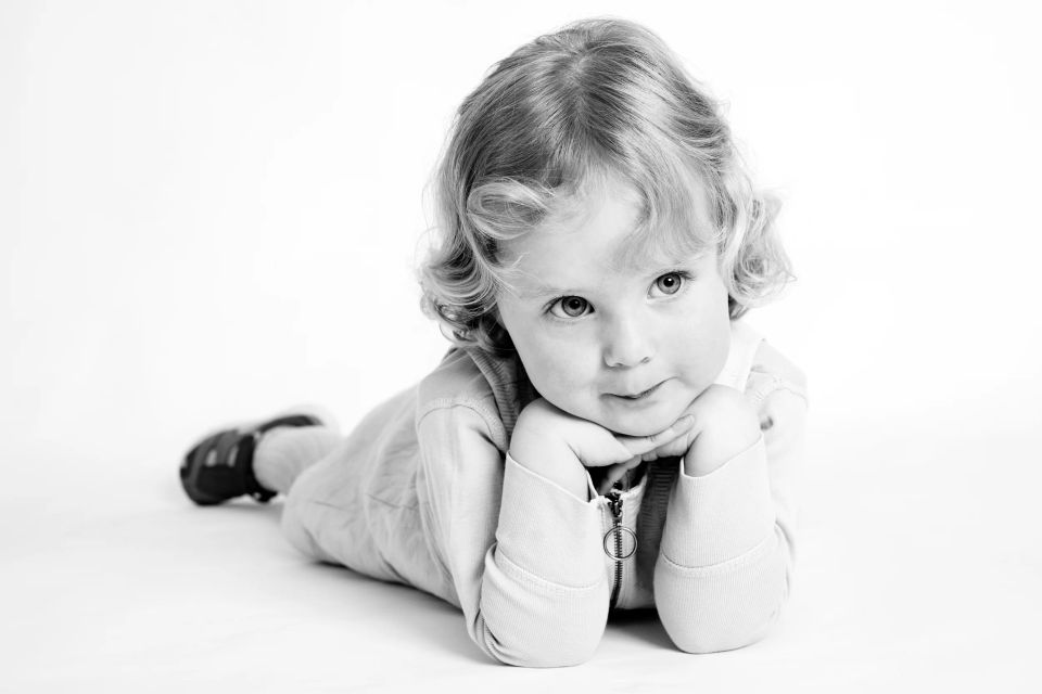 Sort-hvidt børneportræt af barn liggende på maven med hænderne under hagen, der ser direkte ind i kameraet.
