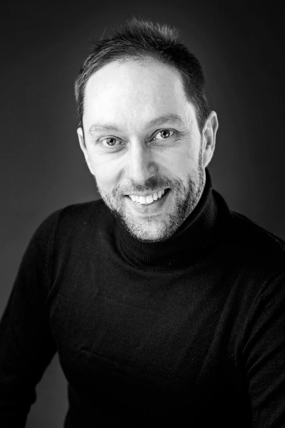 Sort-hvidt portrætfoto af smilende mand med kort skæg og mørk rullekrave mod mørk baggrund.