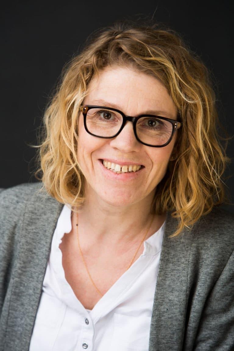 Glad kvinde med briller og lyst krøllet hår smiler varmt i et professionelt portrætfoto med blød belysning.