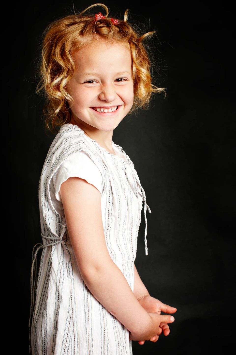 Autentisk børnefotografi af glad pige med krøller og hvid kjole, der smiler begejstret mod sort baggrund.