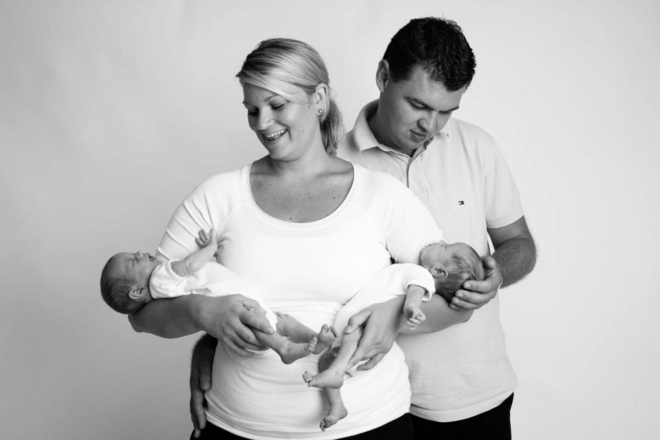 Sort/hvid babyfotografering af smilende mor og far der holder deres nyfødte tvillinger, tydelig glæde i billedet.
