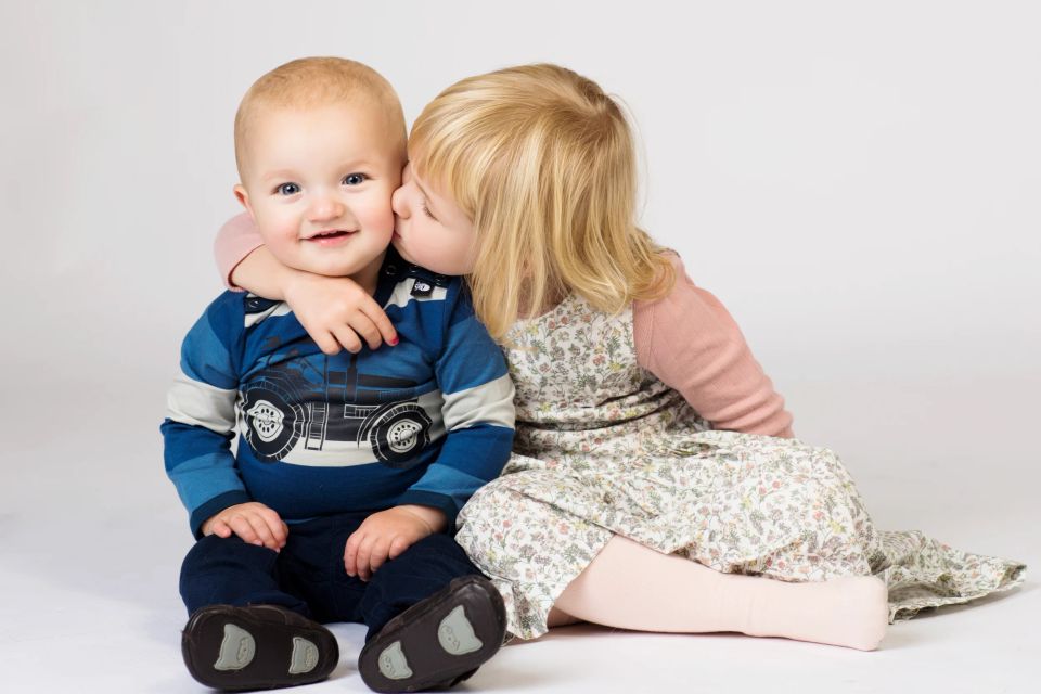 Børneportræt: Større pige giver en lille dreng et kærligt kys på kinden. De sidder tæt sammen, og drengen smiler.