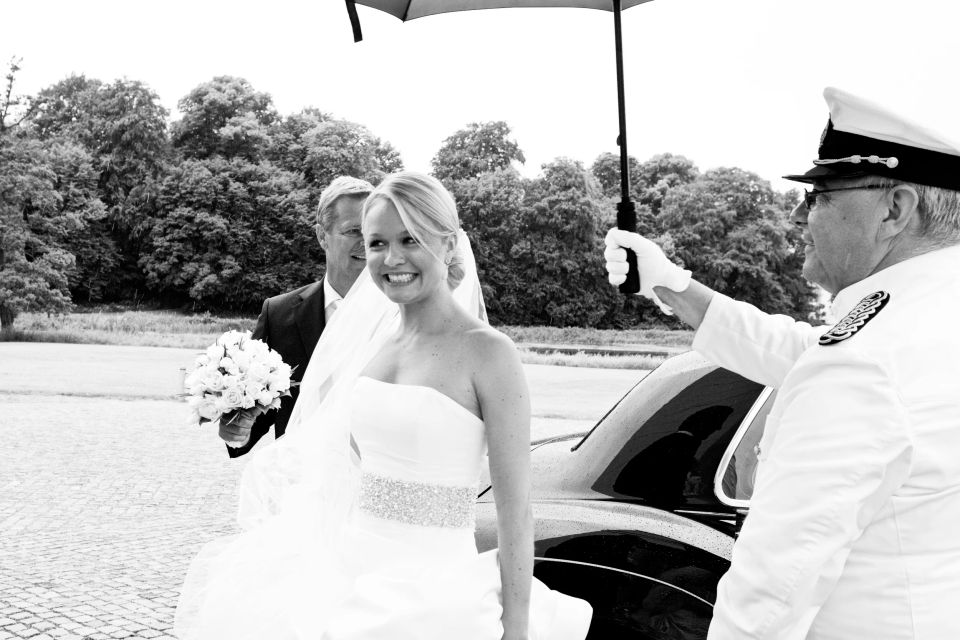 Glad brud smilende under blå parasol, gom ses bagved med brudebuket. Bryllupsfoto i grønne omgivelser.