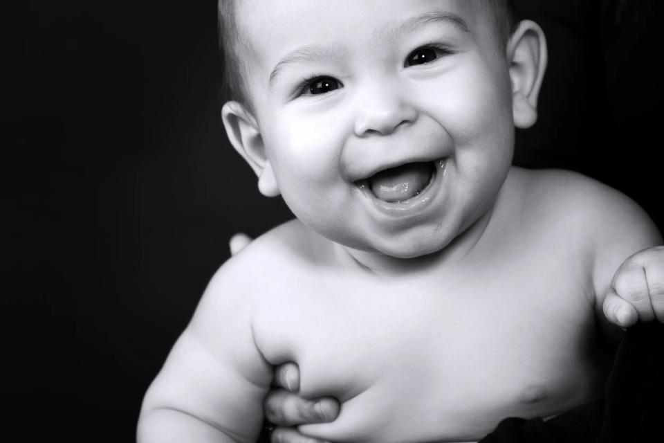 Nærbillede af glad baby med stort smil i sort/hvid fotografering, holder armene op foran brystet. Autentisk babyfoto.