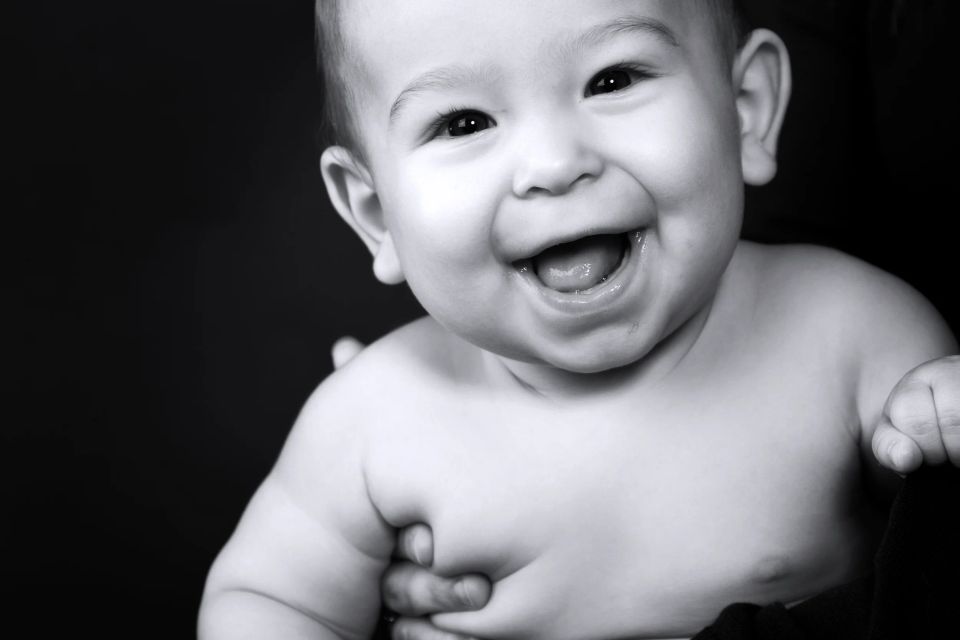 Nærbillede af glad baby med stort smil i sort/hvid fotografering, holder armene op foran brystet. Autentisk babyfoto.