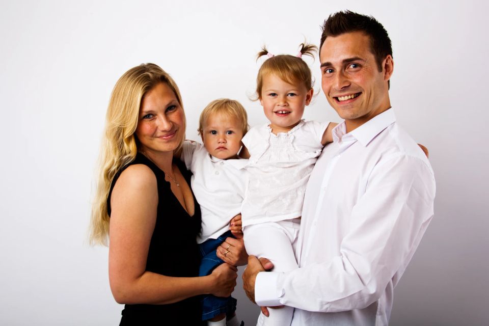 Familieportræt med mor, far og to glade småbørn. Børnene smiler og er i fokus mod hvid baggrund. Familien er let belyst.