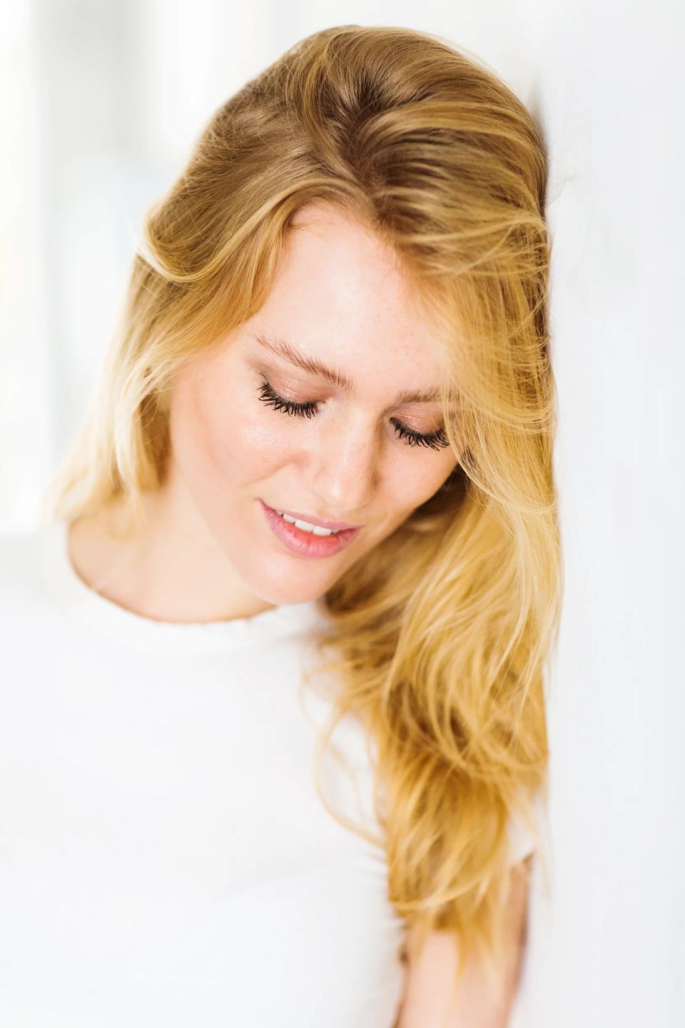 Portrætfoto af ung blond kvinde med lukkede øjne og et venligt smil i blødt, neutralt baggrundslys.