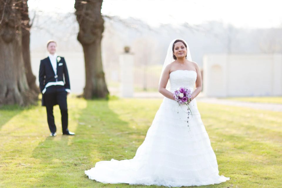 Bryllupsfotografering i sort/hvid af en brud i hvid kjole med brudebuket, stående på en græsplæne. Brudgom i baggrunden.