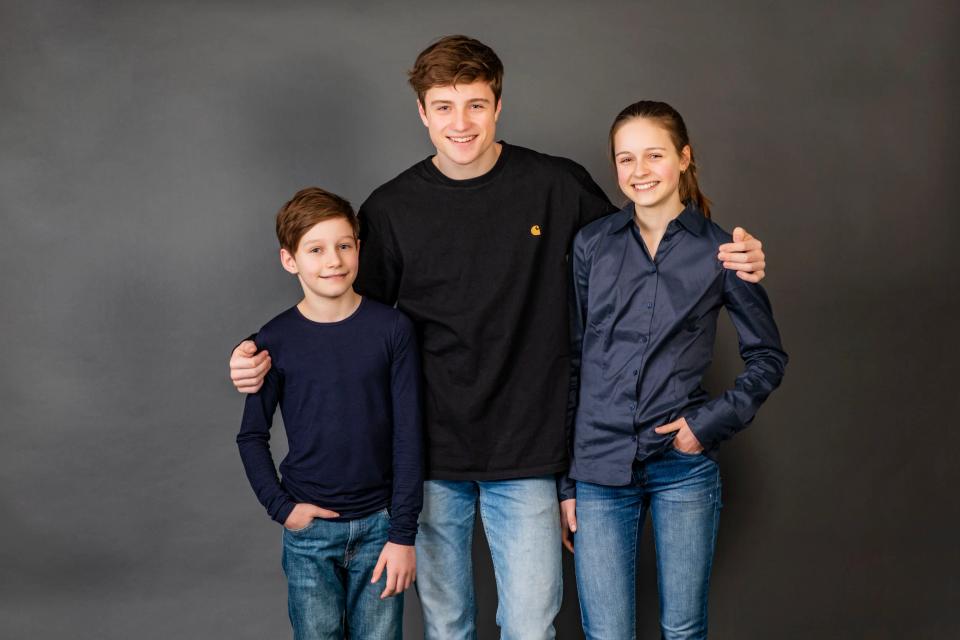 Familiefoto af tre smilende søskende, en dreng, en pige og en ung mand, der omfavner hinanden på en grå baggrund. De er kl...