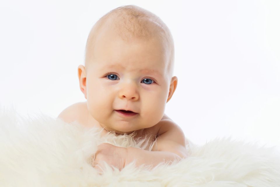 Blåøjet baby i babyfotografering, der ligger på hvidt lammefælde med mildt smil. Naturligt portræt.
