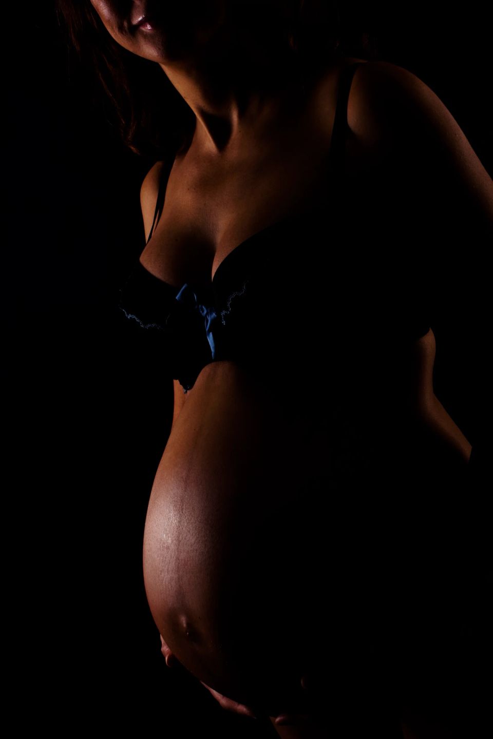 Gravid kvinde i mørkt undertøj smukt belyst fra siden, der fremhæver hendes gravide mave i dæmpet lys.