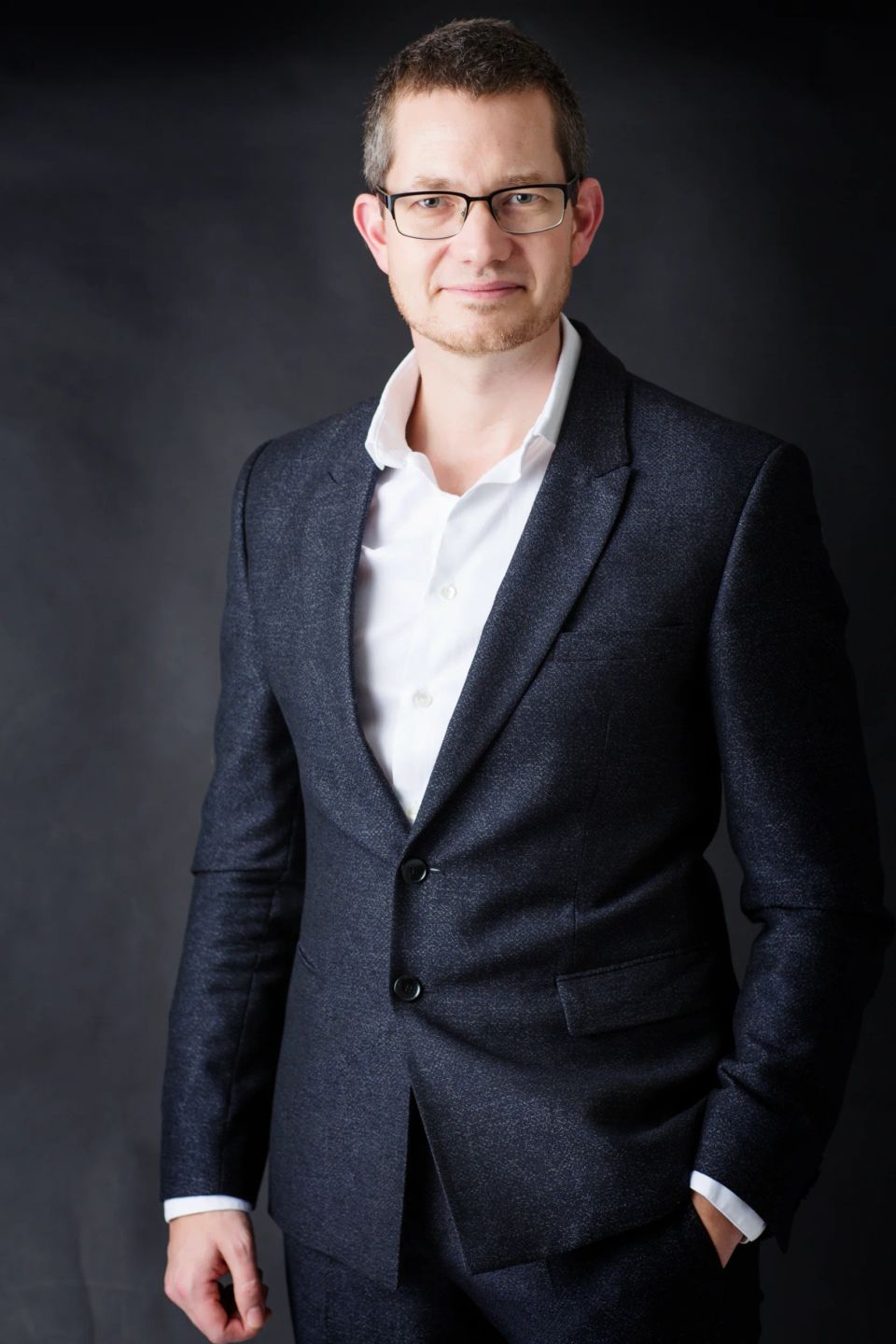 Portrætfoto af mand i gråt jakkesæt med hvid skjorte og briller, stående foran en mørk baggrund med et selvsikkert blik.