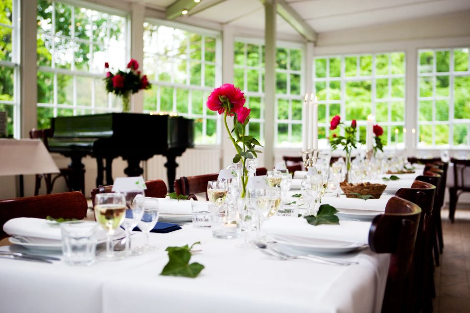 Bryllupslokale fotografering. Langt hvidt dækket bord med lyserøde blomster og levende lys ved store vinduer mod grønt.