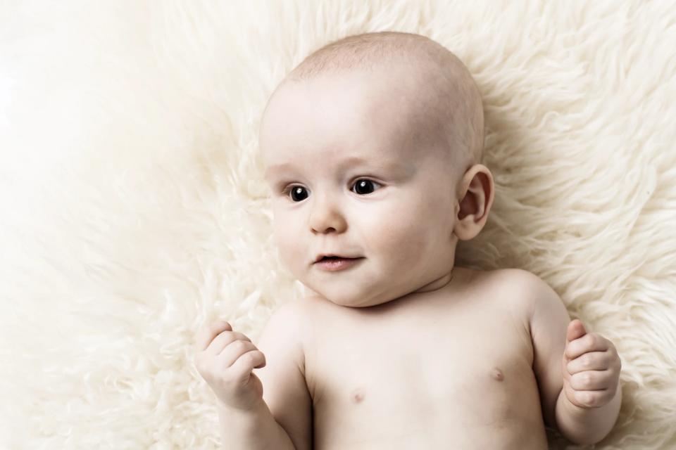 Babyfotografering af spædbarn med åbne øjne, liggende på lyst, blødt tæppe med et nysgerrigt udtryk.