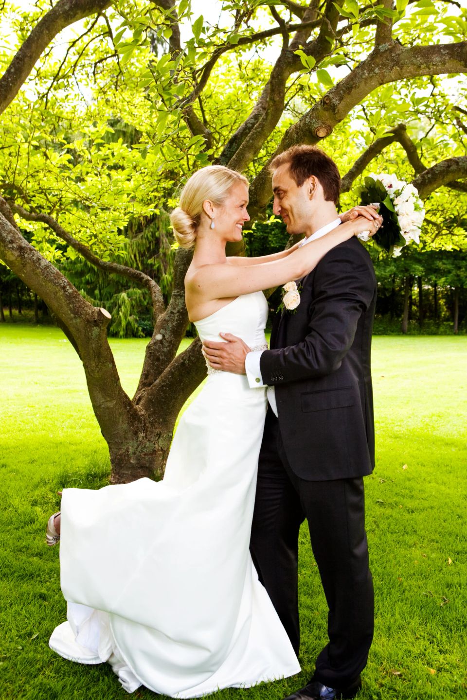 Bryllupsfotografering: Smilende brudepar i omfavnelse under et træ med grønne blade på en sommerdag.