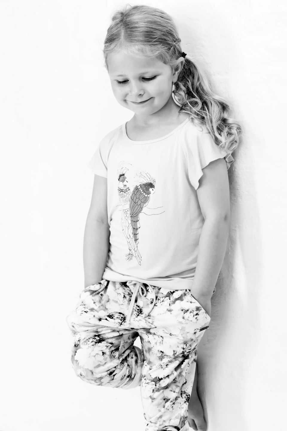 Sort-hvid fotografering af smilende, legende pige i Virum med hænderne i lommen op ad en væg. Et børnebillede i naturligt ...