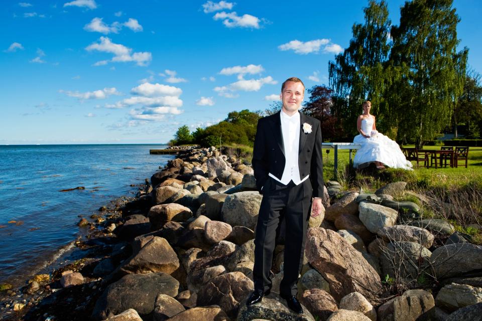 Bryllupsfoto af brudgom stående på sten ved vandet, med bruden siddende i baggrunden på en grøn sommerdag.