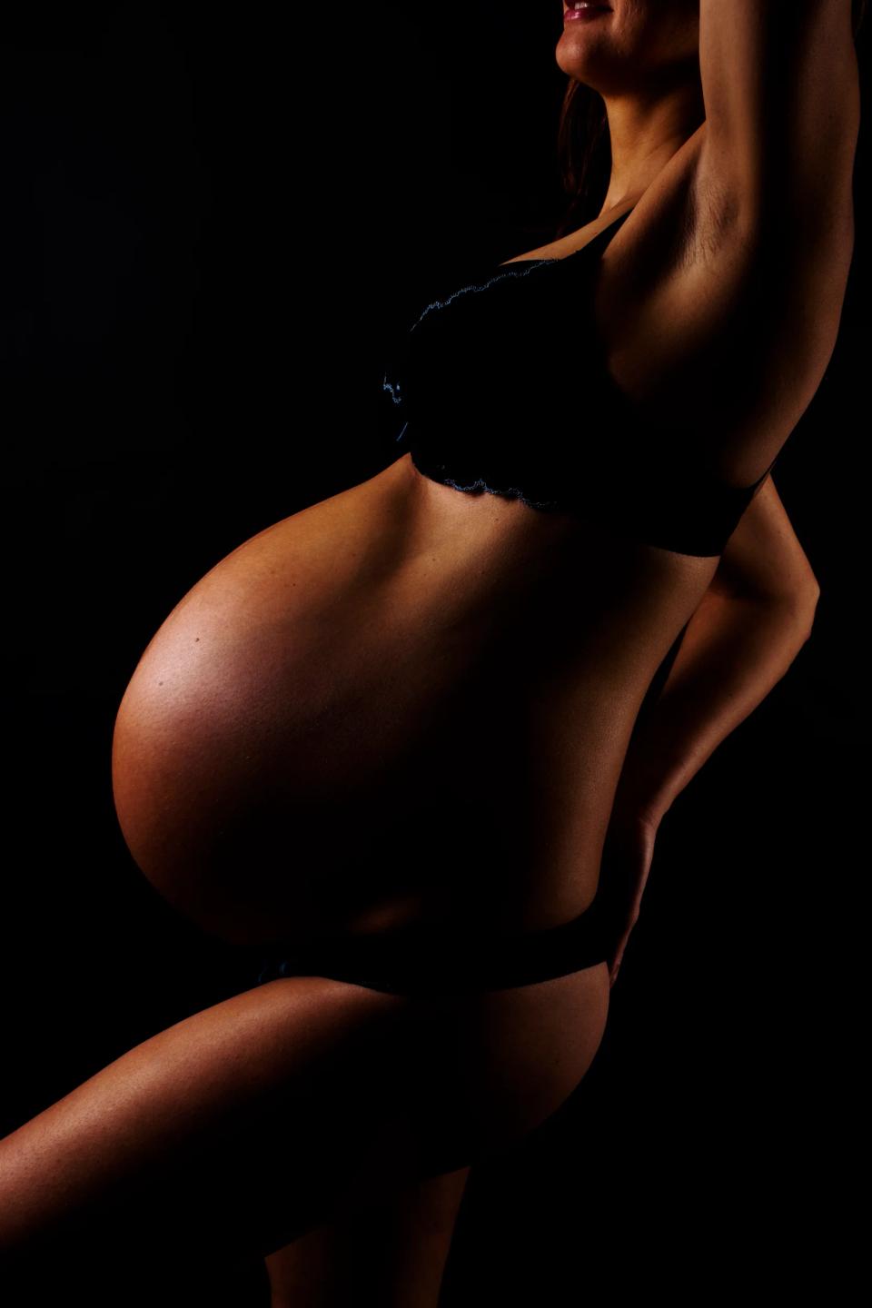 Stemningsfuldt graviditetsfoto: En gravid kvindes silhuet fremhævet af skarpt sidelys, skabende dybe skygger mod mørk bagg...