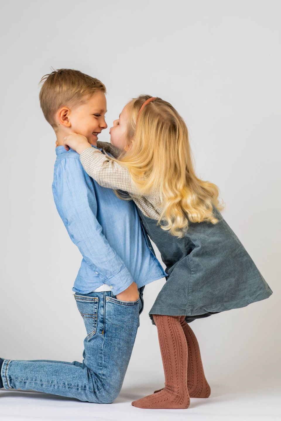 Familiefoto med søskende: Dreng på knæ og pige der knuser ham, begge smiler kærligt. Stemning af glæde og leg.