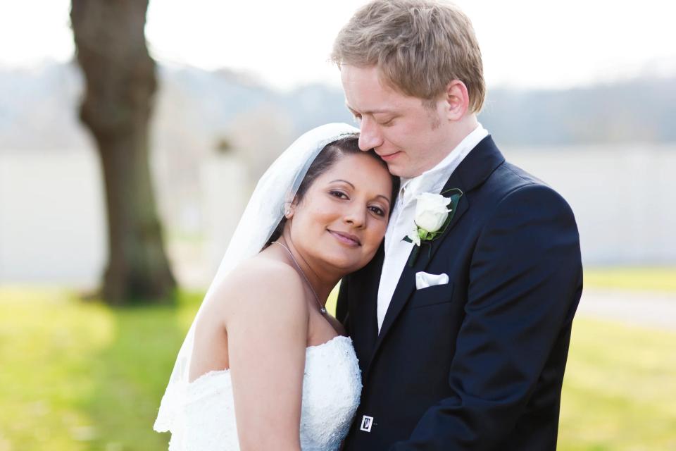 Bryllupsfoto i sorthvid af brudepar i tæt omfavnelse, bruden smiler ind i kameraet, brudgommen kysser hendes pande.