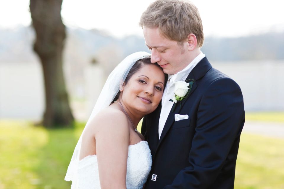 Bryllupsfoto i sorthvid af brudepar i tæt omfavnelse, bruden smiler ind i kameraet, brudgommen kysser hendes pande.