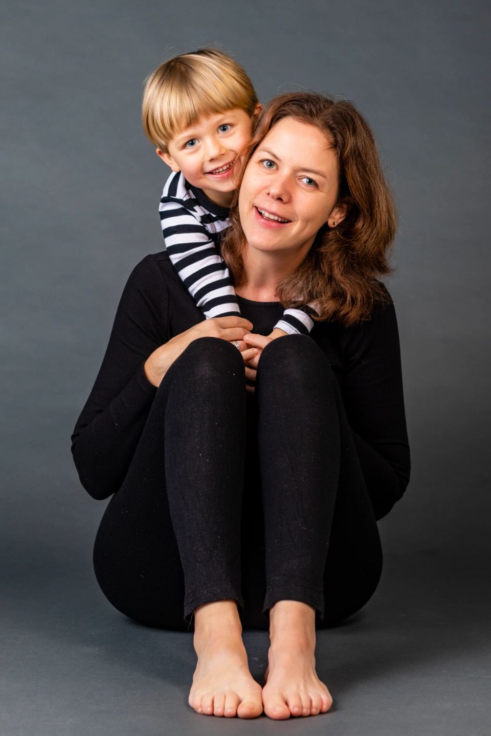 Familiefoto af smilende mor med brune lokker og barn med lyst hår, der kigger frem over hendes skulder, mod grå baggrund.