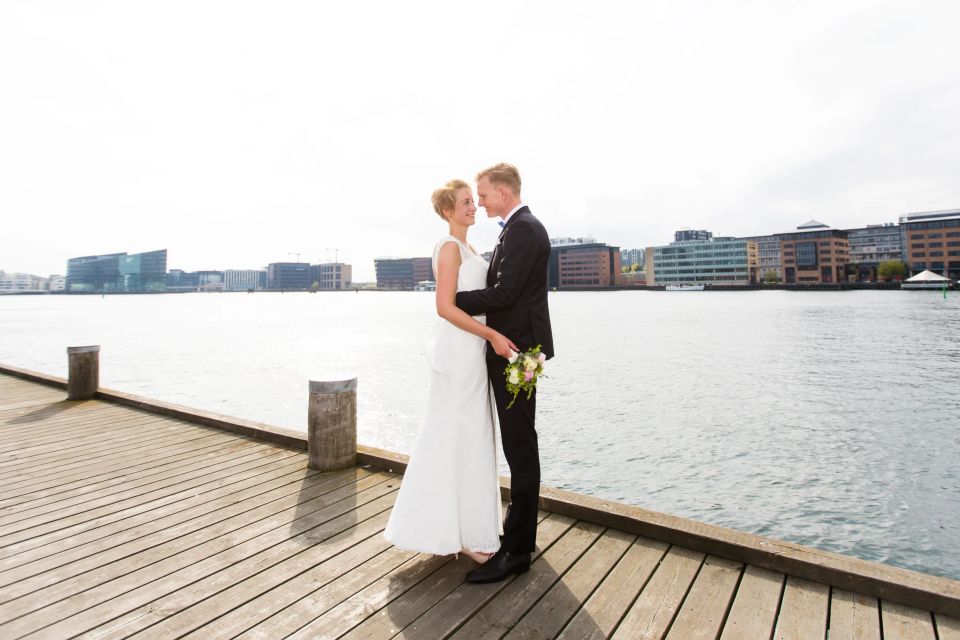 Bryllupsfoto: Et brudepar står tæt omfavnet på en træmolen ved vandet, med bybilledet i baggrunden under dagslys.