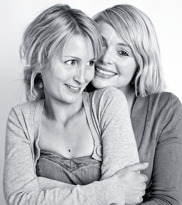Sort/hvid portrætfoto af to glade, blonde kvinder der omfavner hinanden med smil og varme; den ene kigger væk, den anden m...