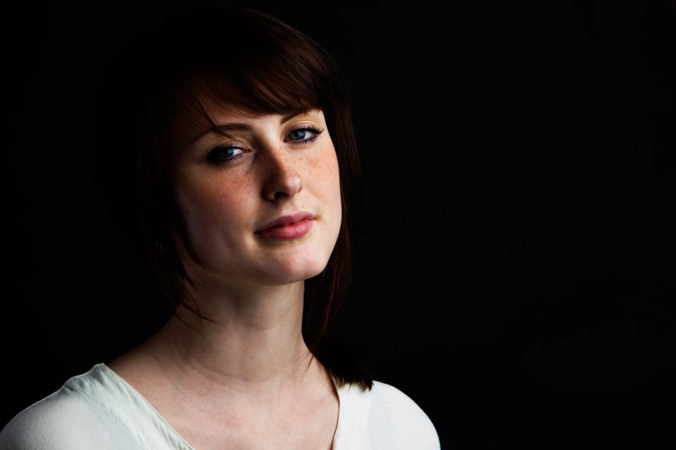 Portrætfoto af kvinde med fregner og brunt hår. Ansigtet er belyst af blødt, naturligt lys mod mørk baggrund.