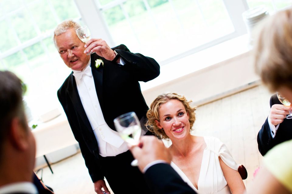 Glad brud smiler under en bryllupsfest, mens en mand i smoking holder en skål op i et naturligt oplyst rum.