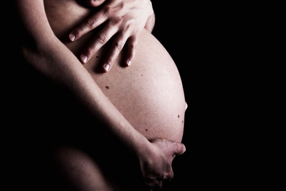 Nærbillede af en gravid mave, holdt blidt af hænderne, mod en mørk baggrund, der skaber fokus på formerne. Gravid fotograf...