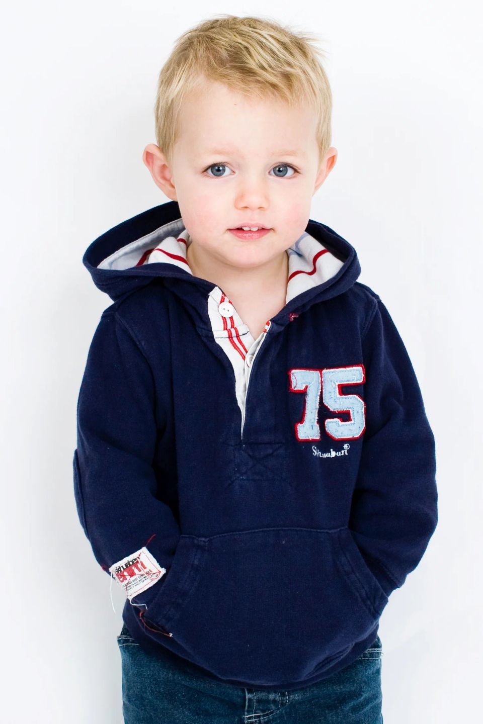 Personligt børnefotografi af en ung lyshåret dreng i mørkeblå hættetrøje stående foran en hvid baggrund.