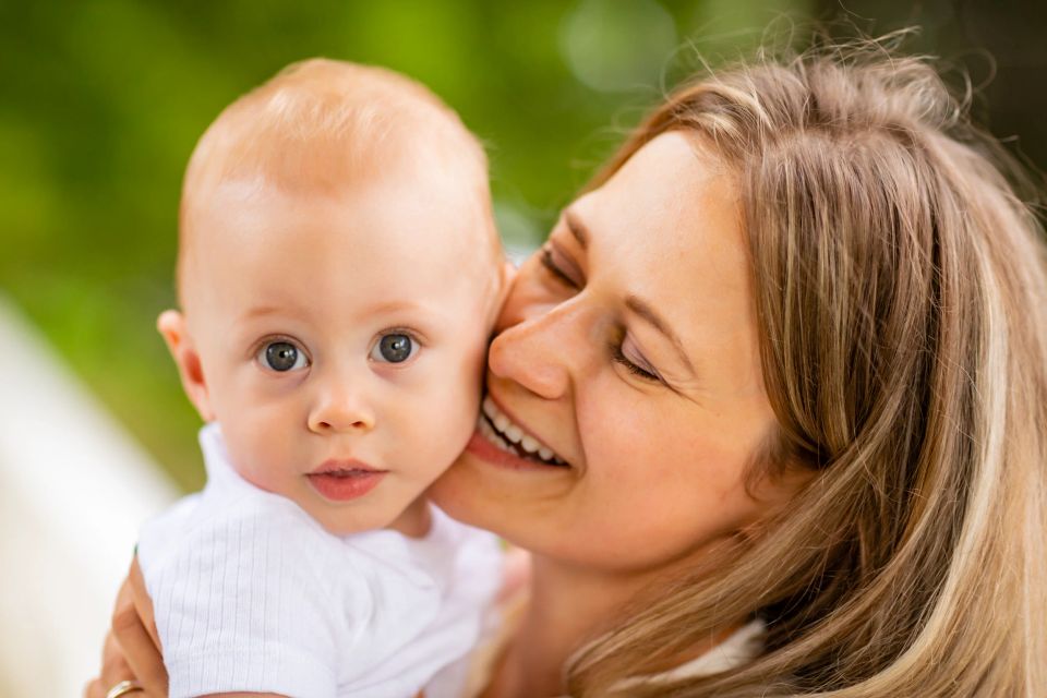 Glad mor kysser sit lyse lille barn på kinden, som kigger nysgerrigt frem. Et hjertevarmt familie foto med blødt lys.