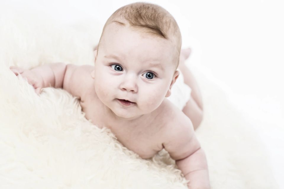 Babyfotografering: Sød baby med åbne øjne, der ligger på maven på et blødt, lyst tæppe og kigger nysgerrigt fremad.