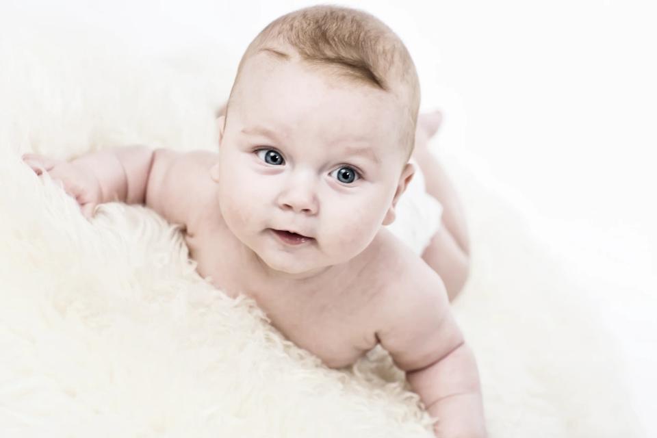 Babyfotografering: Sød baby med åbne øjne, der ligger på maven på et blødt, lyst tæppe og kigger nysgerrigt fremad.