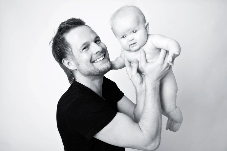 Sort-hvidt babyfoto hvor en smilende far holder sin baby op i luften med blødt, naturligt lys. Stemning af glæde.